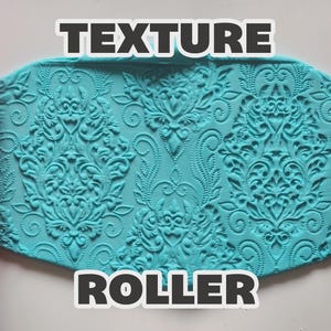 May include: Two red cylindrical texture rollers positioned on the left and right sides of a central piece of light blue clay. The clay features a deeply embossed, intricate damask pattern. The text "TEXTURE ROLLER" is printed in white letters.