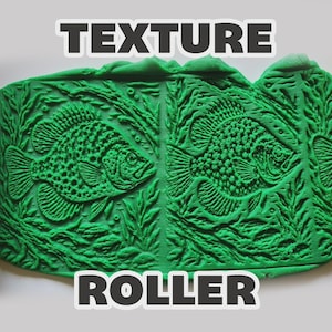 May include: Two yellow cylindrical texture rollers flanking a central green slab of clay. The clay features a repeating embossed pattern of a fish swimming among aquatic plants. The text "TEXTURE ROLLER" is printed in white at the top and bottom.