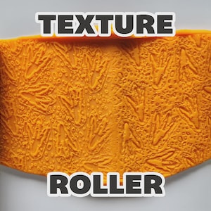 May include: Two light green cylindrical texture rollers flanking a central orange rectangular sheet of material. The surface of the rollers and the orange sheet feature a repeating embossed pattern of three-toed dinosaur footprints and small circular dots. The text "TEXTURE ROLLER" is printed in white with a black outline.