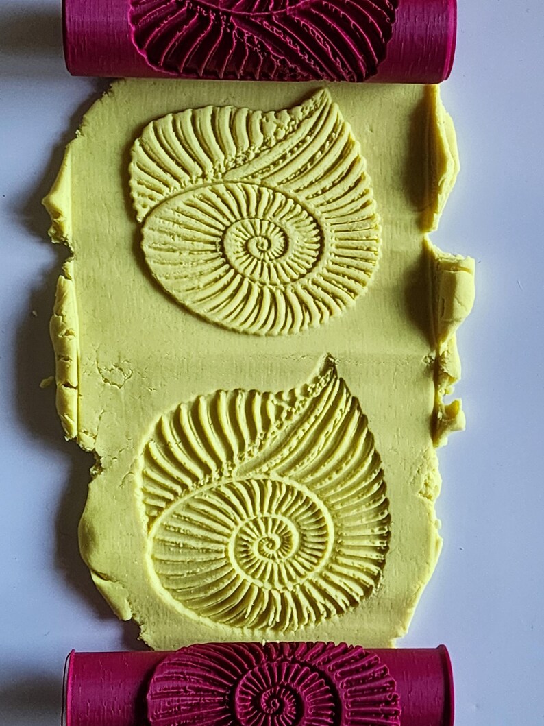 May include: A bright pink cylindrical rolling pin with a deep-set ammonite fossil pattern, shown pressing two identical, detailed spiral shell impressions into a flat, rectangular piece of pale yellow modeling clay or dough.