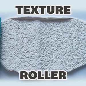 May include: Two bright blue cylindrical texture rollers flanking a central piece of white clay. The clay surface is embossed with a detailed lunar landscape pattern featuring craters and sun-like rays. The text "TEXTURE ROLLER" appears in bold.