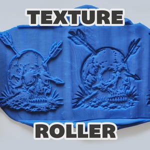 May include: A bright pink cylindrical texture roller featuring a repeating engraved design of a skull pierced by two arrows. The roller is shown pressing into a flat sheet of blue modeling clay, leaving a clear, raised impression of the skull motif.