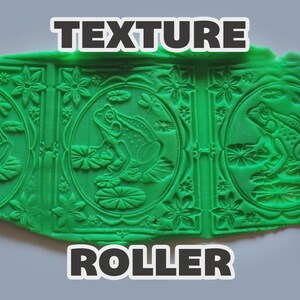 May include: A pink cylindrical texture roller with a repeating pattern of frogs on lily pads, dragonflies, and flowers, shown pressing its design into a flat sheet of bright green clay. The text "TEXTURE ROLLER" is printed in white with a black outline.