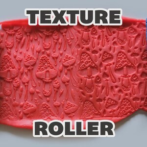 May include: A blue cylindrical texture roller with a repeating mushroom pattern, shown pressing into a flat sheet of red clay. The embossed design features various sizes of mushrooms with dotted caps and stems, creating a consistent surface texture.
