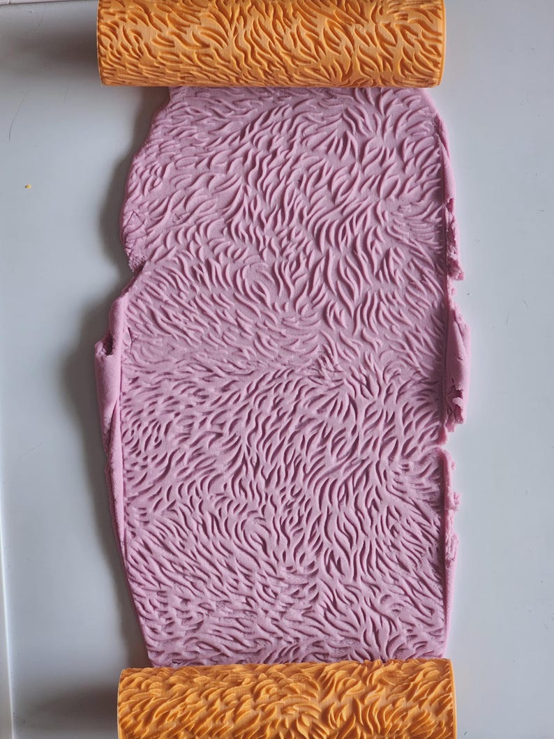 May include: An orange cylindrical rolling pin with a carved, repeating organic texture pattern. The rolling pin is positioned at the top and bottom of a flat, rectangular sheet of light pink clay that has been embossed with the same pattern.