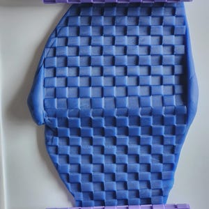 Pu&ograve; includere: Un mattarello cilindrico color viola chiaro con un motivo a griglia quadrata in rilievo, posizionato sopra e sotto un pezzo piatto di pasta modellabile blu che presenta la stessa texture a scacchi su tutta la sua superficie.