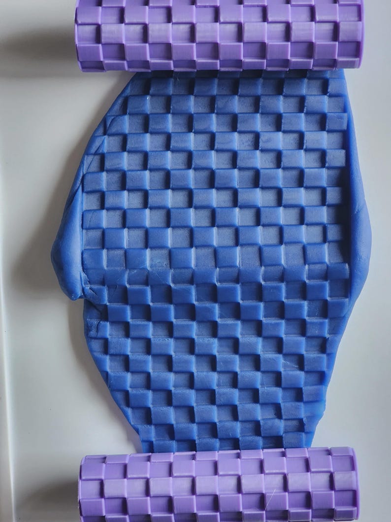 Pu&ograve; includere: Un mattarello cilindrico color viola chiaro con un motivo a griglia quadrata in rilievo, posizionato sopra e sotto un pezzo piatto di pasta modellabile blu che presenta la stessa texture a scacchi su tutta la sua superficie.