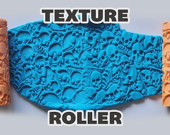 May include: An orange cylindrical texture roller with a repeating skull and bone pattern, shown pressing into a flat piece of bright blue clay. The text "TEXTURE ROLLER" is printed in bold white letters with a dark outline above and below the clay.
