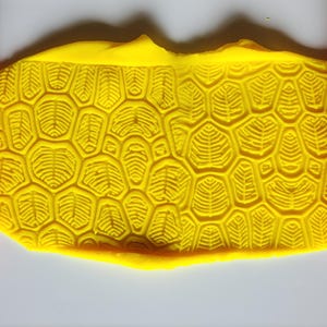 May include: A bright yellow slab of clay featuring a deeply embossed turtle shell pattern, flanked on both sides by orange cylindrical rollers that display the same intricate, repeating geometric texture used to create the impression.