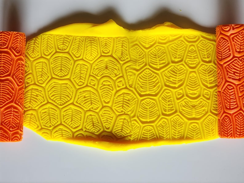 May include: A bright yellow slab of clay featuring a deeply embossed turtle shell pattern, flanked on both sides by orange cylindrical rollers that display the same intricate, repeating geometric texture used to create the impression.