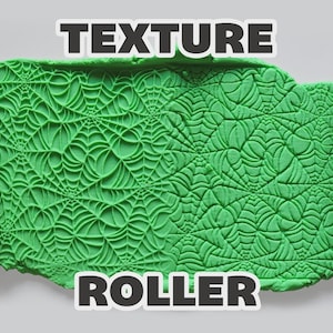 May include: Two magenta cylindrical texture rollers with a spider web pattern positioned on either side of a green clay slab imprinted with the same spider web design. The text "TEXTURE ROLLER" is displayed in white letters at the top and bottom.