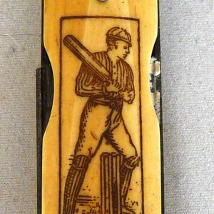 May include: Antique cricket bat-shaped pocket knife. The handle is a light tan color with a textured grip. A rectangular image of a cricket player is on the blade. The text "RD 172718" is visible. The knife is closed.