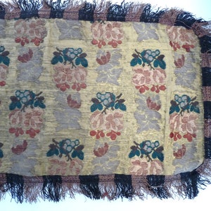 May include: A close-up of a fabric with a repeating pattern of pink flowers and green leaves. The fabric has a black and brown fringe border.