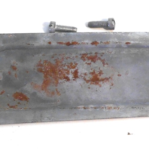 May include: A rusty metal oval-shaped cover with two screw holes on each side. The cover has a raised oval in the center and is slightly curved.