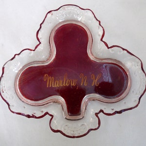 May include: A red glass trinket dish with a clear glass rim. The dish is shaped like a clover and has the inscription "Marlow, N.H." in gold lettering.