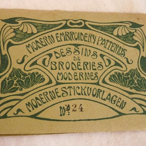 May include: Vintage embroidery pattern booklet with green Art Nouveau style lettering and illustrations on a tan background. The text reads "Modern Embroidery Patterns" in English, French, and Dutch. Number 24 is printed at the bottom.