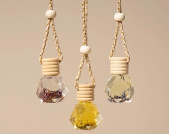 Puede incluir: Tres frascos de vidrio para ambientador con facetas geométricas, cada uno con un tapón de madera clara y suspendidos por un cordón trenzado con una cuenta de madera. Los frascos contienen líquido y pétalos de flores secas.