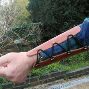 May include: A brown leather arm guard with black straps and metal buckles. The guard is worn on the forearm and is secured with a series of straps and buckles.