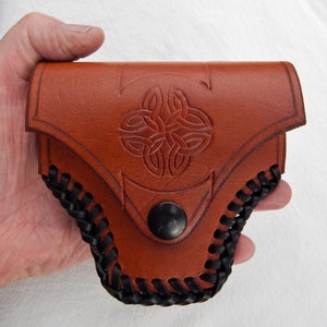 May include: A brown leather pouch with a Celtic knot design embossed on the front. The pouch has a black braided trim and a black snap closure. The pouch is held in a hand.