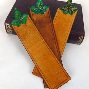 3 Green, cut out leaf leather bookmarks resting on a navy book. Showing the shade differances between these individually handmade, bookmarks in vegetable tanned leather.