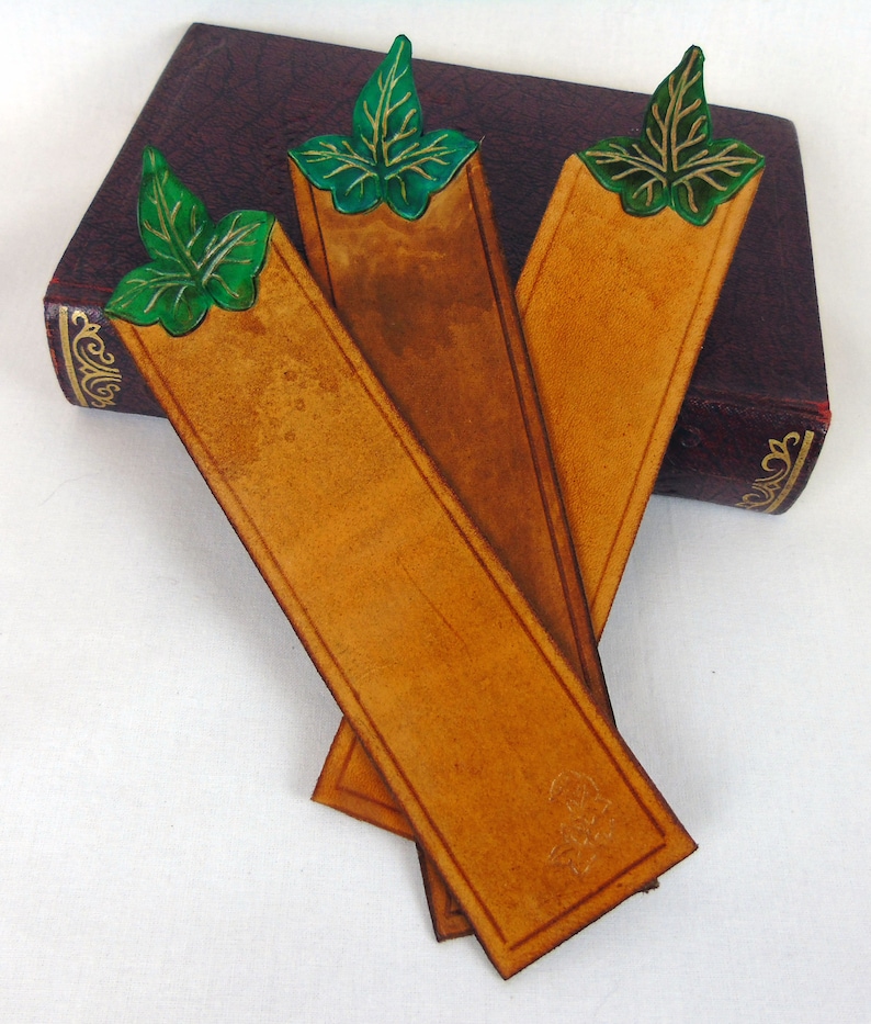 3 Green, cut out leaf leather bookmarks resting on a navy book. Showing the shade differances between these individually handmade, bookmarks in vegetable tanned leather.