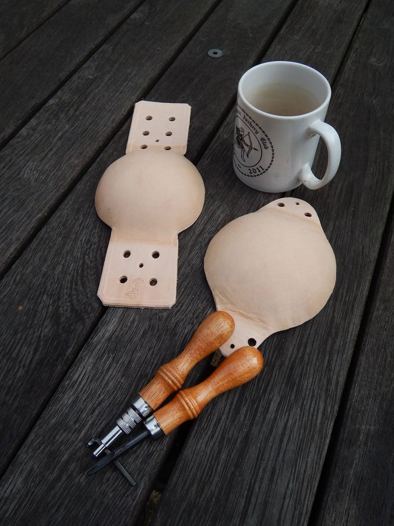 Domed leather bell muffle and seperate overlay on a dark workbench with two of the hand tools used (with a mug of tea for scale)