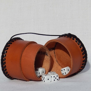May include: Two brown leather dice cups with black stitching and a black cord connecting them. Five white dice with black dots are spilling out of the cups.