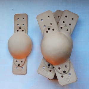 May include: Four tan leather knee pads with a rounded shape and multiple holes. The knee pads are stacked on top of each other.