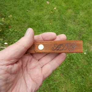 Nubuck tan leather belt clip with brassed trigger clip personalised with ADG, shown held in hand with grass in background