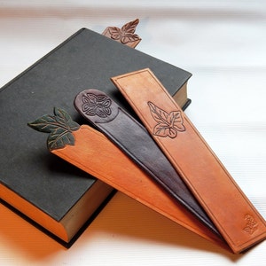 Selection of three different handmade leather bookmarks resting on a navy book.  Left to right, a green, cut out leaf on top, a round celtic knot on a domed top and a rectangular simple one with an embossed leaf in the body.  Tactile and durable.