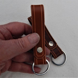 May include: Two brown leather keychains with a brown and orange striped design. Each keychain has a silver metal ring and a silver metal button.