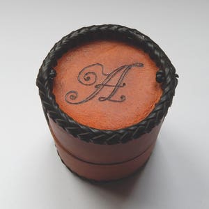Hand made circular leather pot with a snug fitting lid that can be used as a second pot when open.  Vegetable tanned leather with a plait like black thong stitching around the base and top secured, show with A burnt on to the lid.
