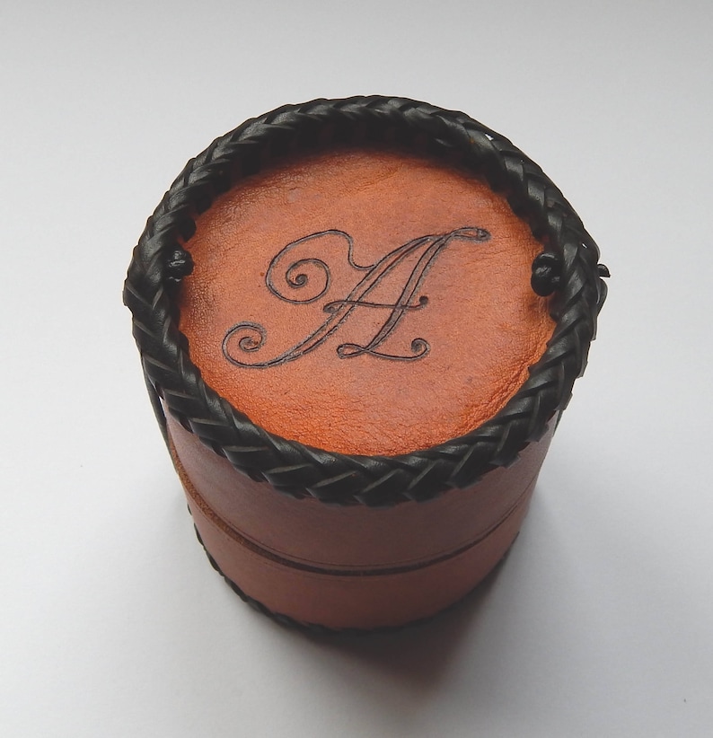 Hand made circular leather pot with a snug fitting lid that can be used as a second pot when open.  Vegetable tanned leather with a plait like black thong stitching around the base and top secured, show with A burnt on to the lid.