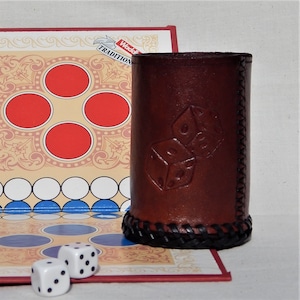 May include: A brown leather dice cup with a braided trim. The cup has two dice embossed on the front, one showing a five and the other a six. The cup is sitting on a game board with red, white, and blue squares.
