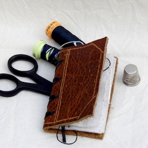 Neat leather covered needle case with four white felt pages for your pins, needles etc. in a variety of different leathers and colours.  Decorated with incised lines to resemble a bound book with a thong stitched spine.  Shown with sewing accessories