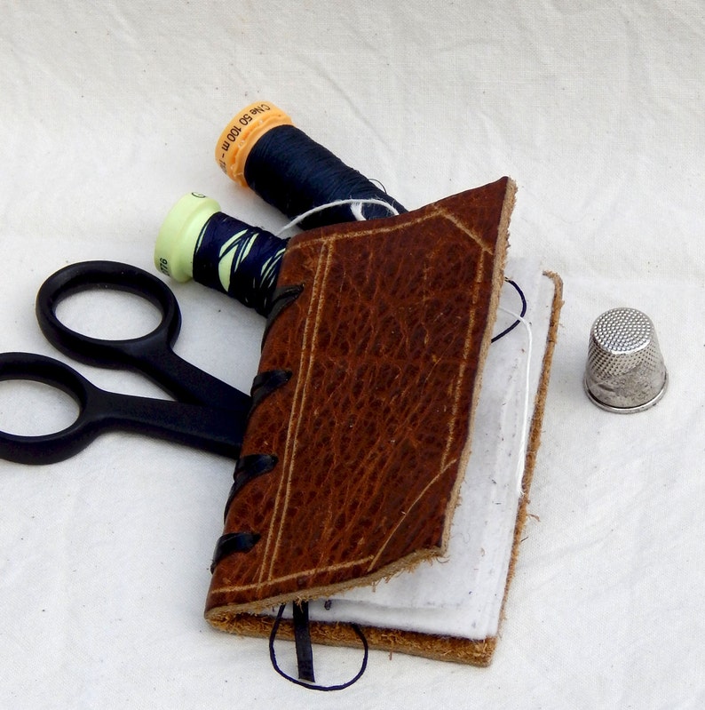 Neat leather covered needle case with four white felt pages for your pins, needles etc. in a variety of different leathers and colours.  Decorated with incised lines to resemble a bound book with a thong stitched spine.  Shown with sewing accessories