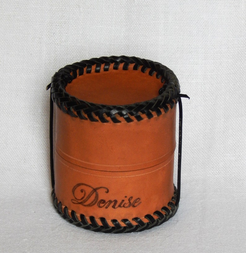 Circular leather pot showing optional personalisation, here with the name Denise burnt in near the base.  Vegetable tanned leather with a plait like thonging around the base and top, thong is twisted beneath to keep it closed.