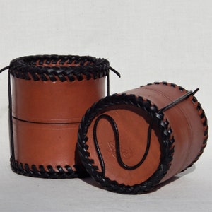 2 circular leather pots with snug fitting lids that can be used as a second pot.  Vegetable tanned leather with a plait like thonging around the base and top, 1 
 upright, closed, one showing how the thong is twisted beneath to keep it closed.