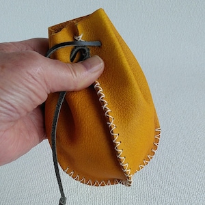 May include: A small, yellow leather pouch with a white stitched border and a black drawstring. The pouch is held in a hand.