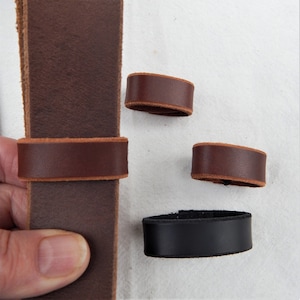 Replacement cut-to-fit belt loops in brown and black leather, here shown made into belt loops.  One brown one fitted onto a belt on the left, held in a hand.  Two brown and one black on the right in different sizes on a neutral backgroud.