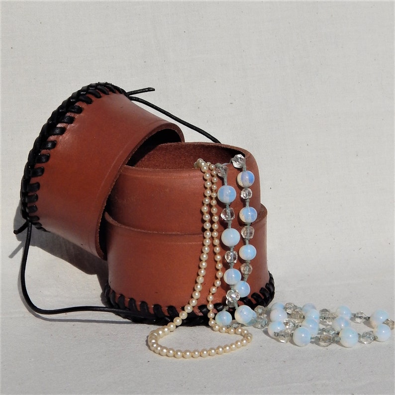A circular leather pot open with necklaces (not included) lid that can be used as a second pot.  Vegetable tanned leather with a plait like thonging around the base and top.