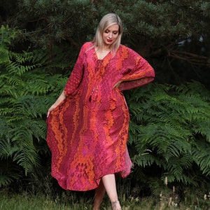 May include: A flowing, knee-length dress with a vibrant snakeskin pattern in shades of red, pink, and orange. The dress features a V-neckline with a tassel tie and loose, three-quarter sleeves. The model is standing in a grassy area with ferns and trees in the background.