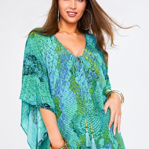 May include: A teal and green patterned kaftan dress with a V-neckline and tie closure. The dress features a snake-skin design and wide sleeves. The model is wearing gold hoop earrings and gold bracelets. The dress has a tassel detail.