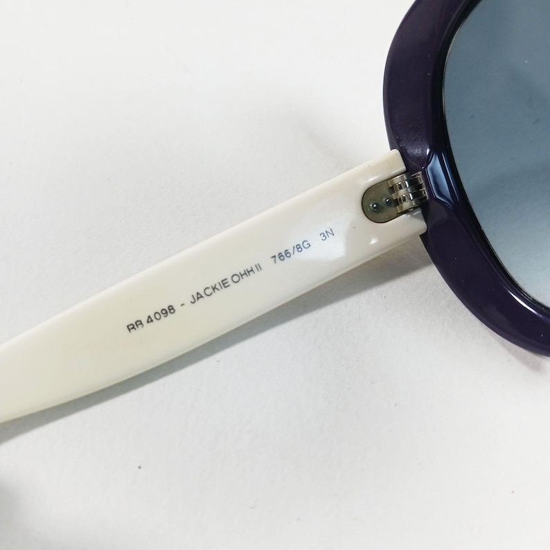 Peut inclure: Gros plan sur des lunettes de soleil avec une monture violette et une branche blanche. Le texte "RA 4098 - JACKIE OHH II 766/8G 3N" est imprim&eacute; sur la branche. Les verres sont bleus d&eacute;grad&eacute;s.