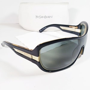 May include: Black sunglasses with a wrap-around frame and dark tinted lenses. The temples feature gold-tone accents with the Yves Saint Laurent logo engraved on the side. A white protective case with the Yves Saint Laurent brand name is included.