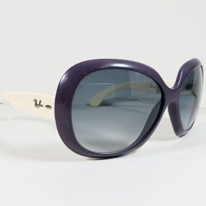 Peut inclure: Lunettes de soleil surdimensionn&eacute;es avec une monture violet fonc&eacute; et des verres d&eacute;grad&eacute;s. Les branches sont blanches avec le logo Ray-Ban. Les lunettes ont une forme arrondie et f&eacute;minine.