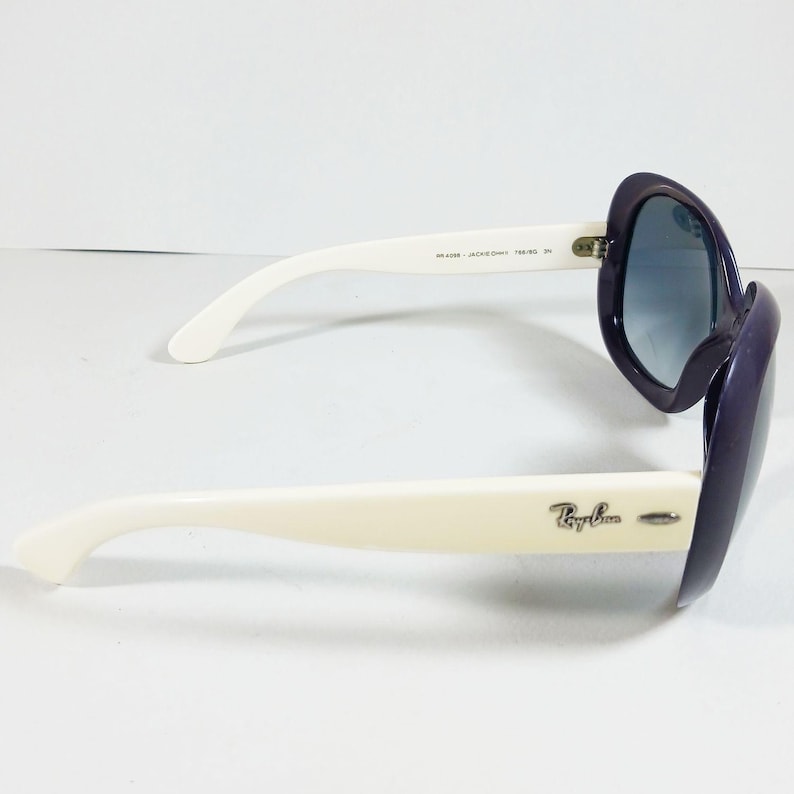 Peut inclure: Lunettes de soleil Ray-Ban violettes et blanches, avec une monture arrondie et des verres d&eacute;grad&eacute;s. Les lunettes ont des branches blanches avec le logo Ray-Ban et une monture violet fonc&eacute;. Les verres ont une teinte bleue.