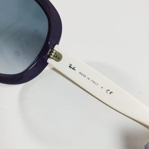 Peut inclure: Gros plan sur des lunettes de soleil Ray-Ban avec une monture violet fonc&eacute; et des verres d&eacute;grad&eacute;s. La branche blanche des lunettes de soleil porte le logo Ray-Ban et l'inscription "MADE IN ITALY". Un accessoire &eacute;l&eacute;gant.