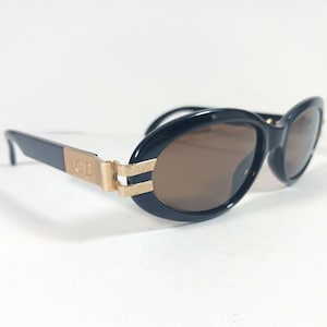 May include: Black oval sunglasses with brown tinted lenses. The temples feature a gold-tone metal accent engraved with the letters CD, connected to the frame by a textured gold-tone metal hinge detail. The frame is made of a smooth black material.