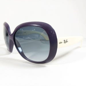 Peut inclure: Lunettes de soleil violettes surdimensionn&eacute;es avec des verres d&eacute;grad&eacute;s et des branches blanc cass&eacute;. Le logo Ray-Ban est visible sur la branche. Les lunettes ont un design r&eacute;tro arrondi.